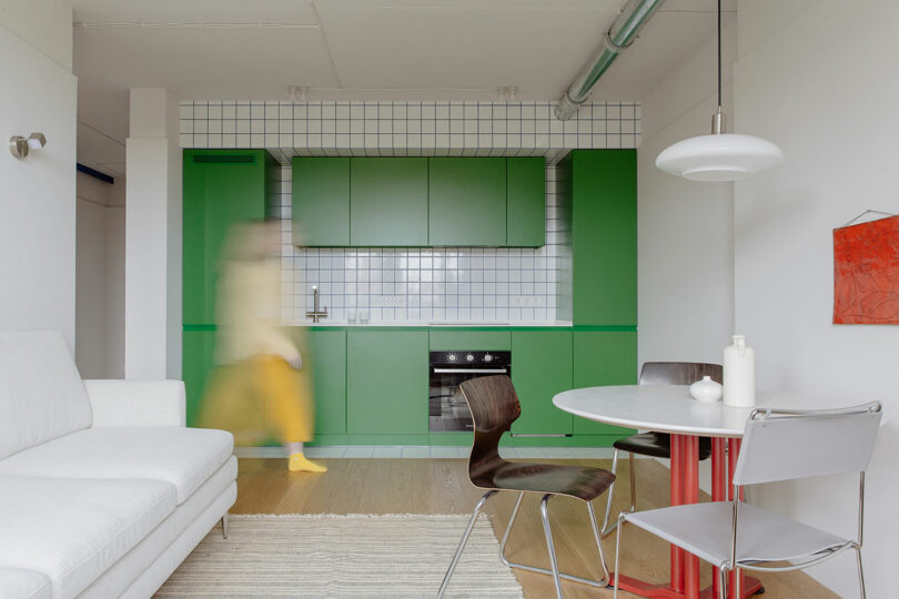 Modern kitchen with green cabinets and white tiled walls. Blurred person in yellow walks by. White sofa and round dining table with chairs in foreground. Red artwork on wall.