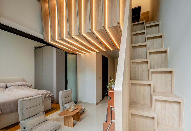 Modern bedroom with a wooden loft ceiling, two chairs, and a small wooden staircase. A bed is on the left and a plant is on the stairs.