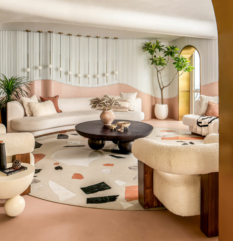 A modern living room featuring a circular rug, plush seating, a round black coffee table, and potted plants. Neutral and earth tones dominate the decor.