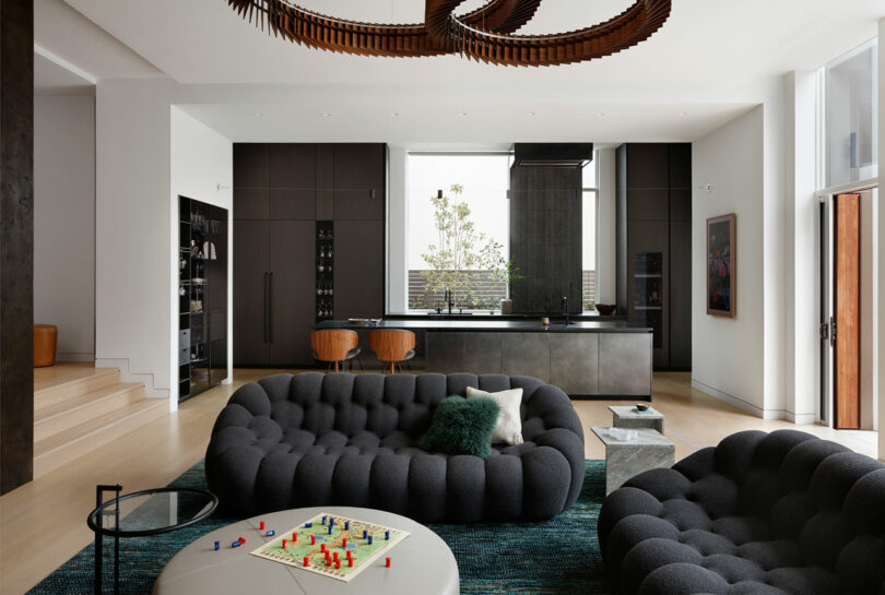 Modern living room with dark gray sofas, a glass coffee table, and a board game. Open kitchen in the background with a large island and bar stools. Contemporary lighting hangs above.