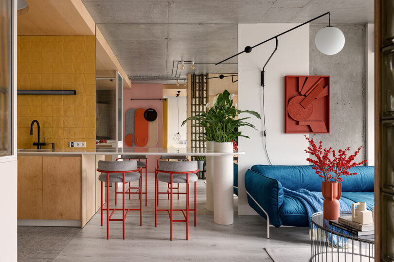 Modern living area with a blue sofa, red accents, and yellow backsplash in the kitchen. A tall plant in a white pot separates spaces.