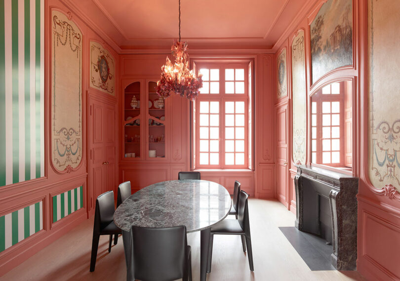 A pink-walled dining room features a marble oval table, six black chairs, a chandelier, a fireplace, and decorative wall panels.