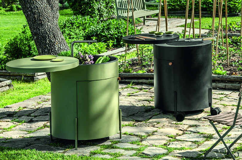 Two cylindrical outdoor grills are set on a stone patio in a garden. One is green, and the other is black. They have hinged lids and are surrounded by lush greenery and garden beds.