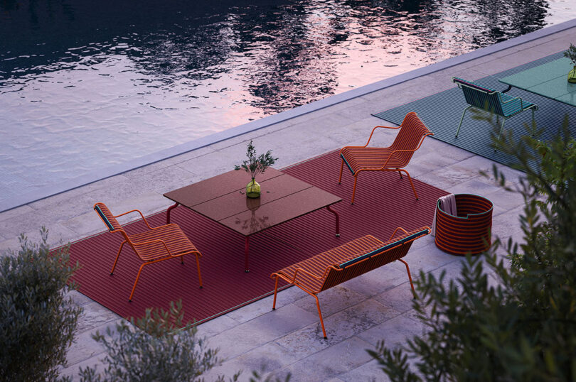 Outdoor seating area by a pool with four orange chairs, two square tables, and a flowerpot on a red mat.