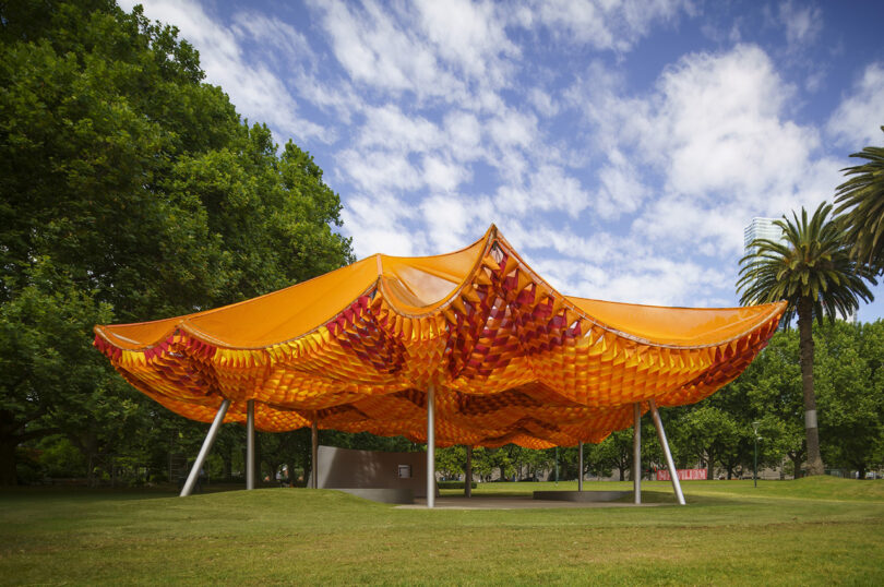 Orange and yellow woven pavilion with metal supports in a park, surrounded by green trees under a partly cloudy sky.