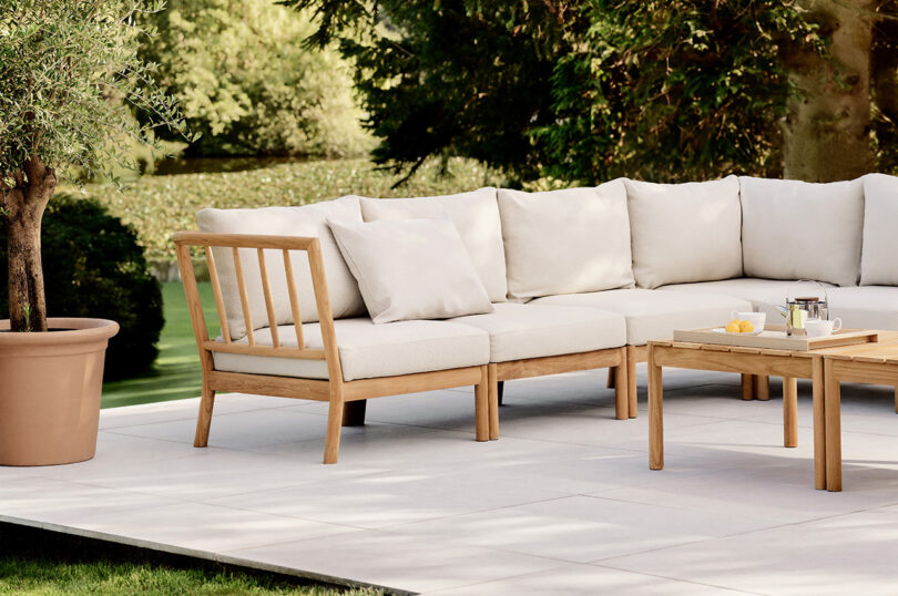 Outdoor seating area with a wooden corner sofa, beige cushions, and a wooden coffee table. The table has a tea set and a plate with fruit. There is a potted plant nearby.