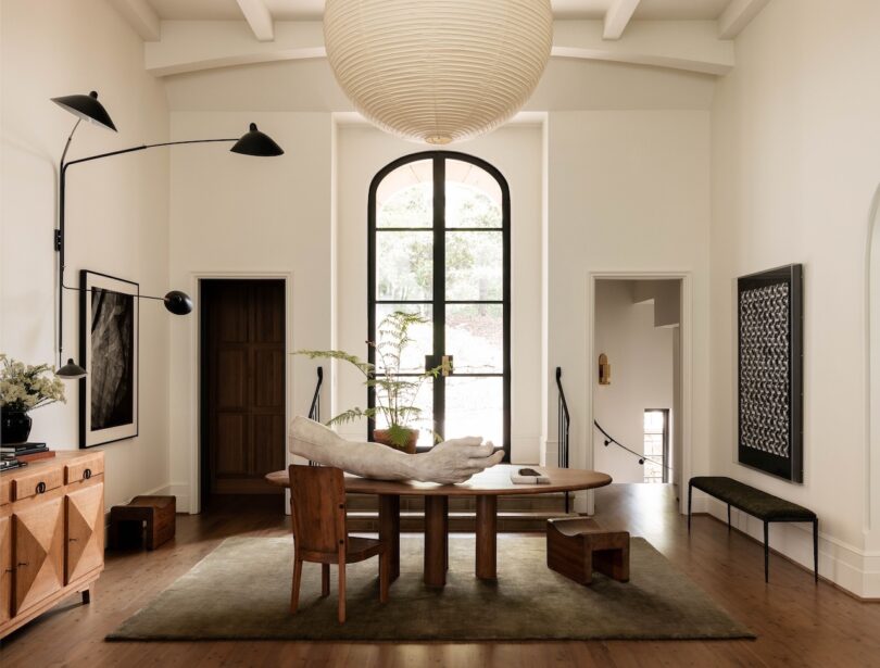 A modern dining room with a round wooden table, chairs, a large window, a paper lantern ceiling light, and various decorations including plants and framed art