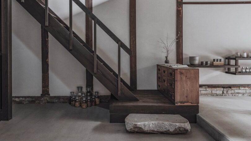 Minimalist interior with a wooden staircase, storage chest, dried branch in a vase, and jars on the floor. Neutral tones and natural materials create a serene atmosphere