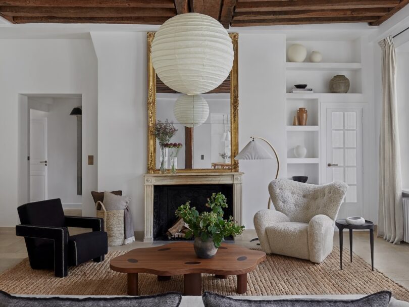 A stylish living room features modern armchairs, a distinctive wooden coffee table, and a large mirror above the fireplace. A spherical paper lantern and decorative items complete the decor