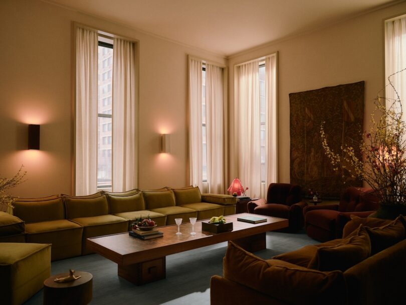 Cozy living room with a long wooden coffee table, green sectional sofa, armchairs, and wall art. Tall windows with sheer curtains allow soft light to illuminate the space