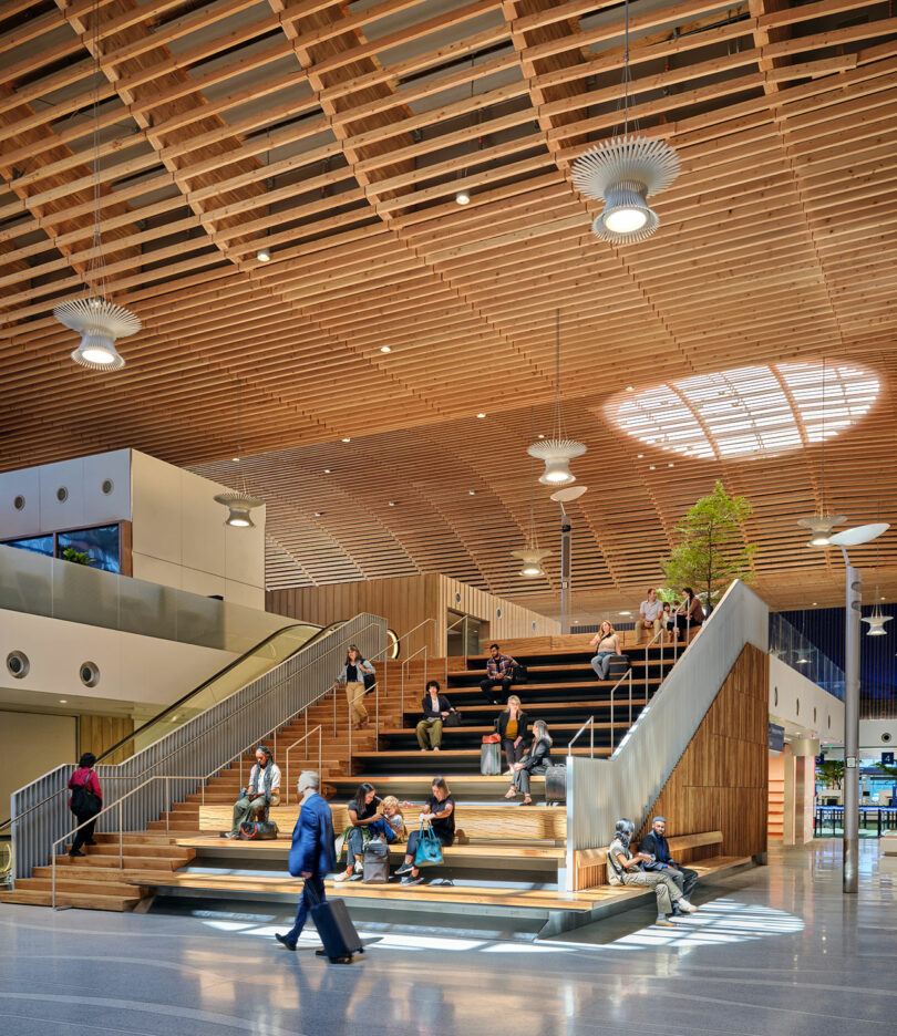 People sitting on a large wooden indoor staircase with a modern, open ceiling design and skylight, reminiscent of ZGF's architectural style. One person walks by with luggage.