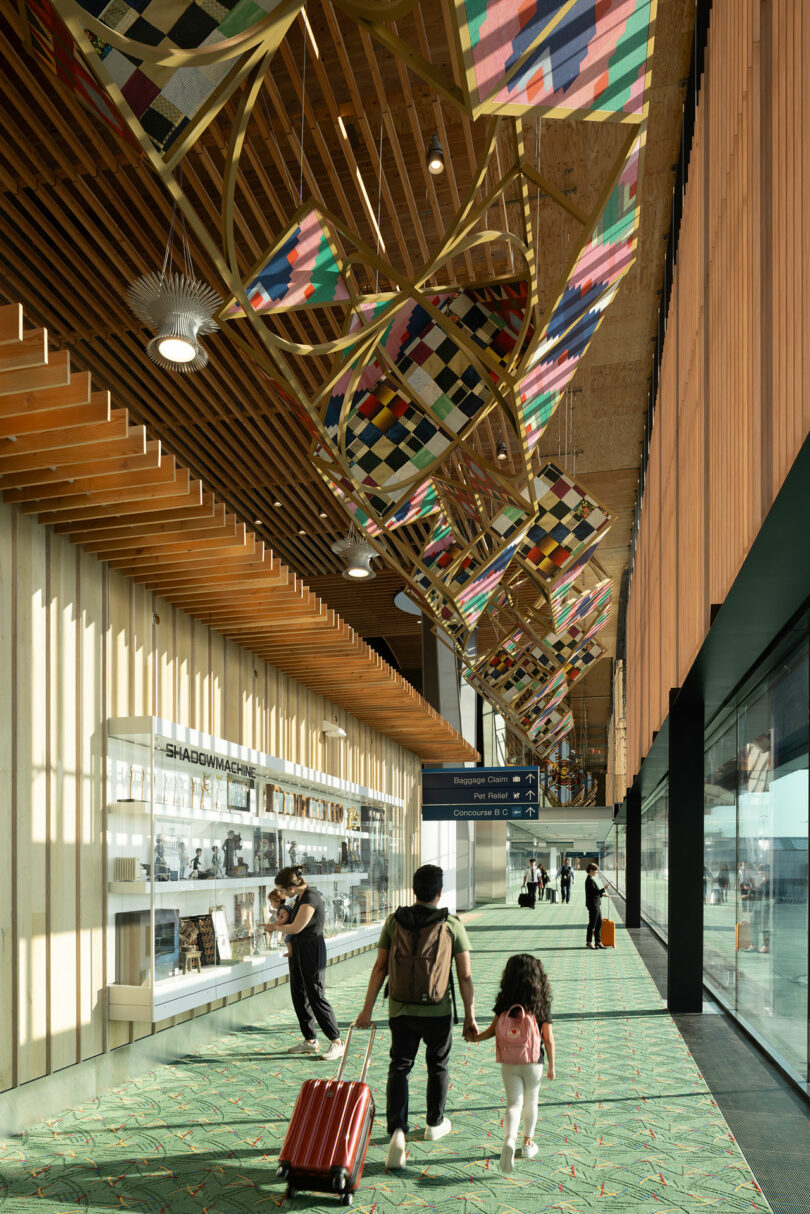 A man and child holding hands walk through an airport terminal designed by ZGF, featuring colorful geometric decor overhead and a display case on the left.