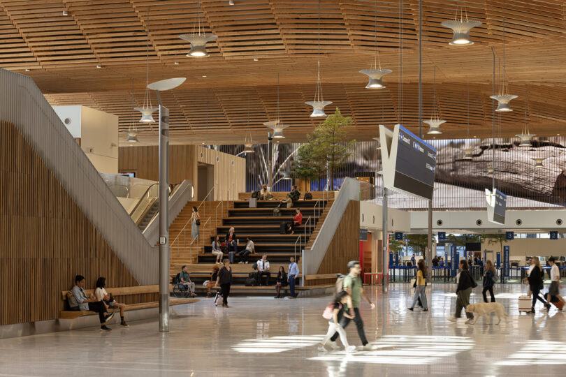 The interior of a large, modern airport terminal designed by ZGF showcases a stunning wooden ceiling. Passengers with luggage stroll and relax on benches and steps, while large screens efficiently display flight information.