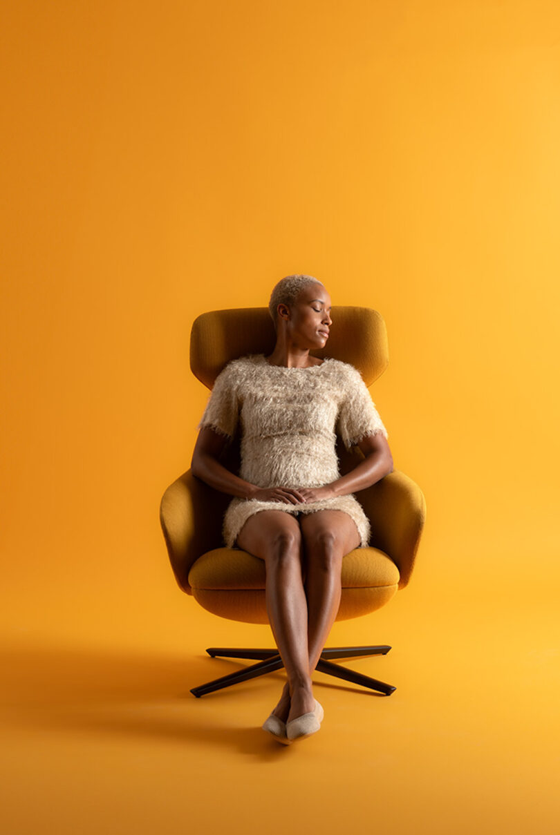 Person seated on a mustard yellow chair against a matching yellow background, wearing a textured beige dress and looking to the side