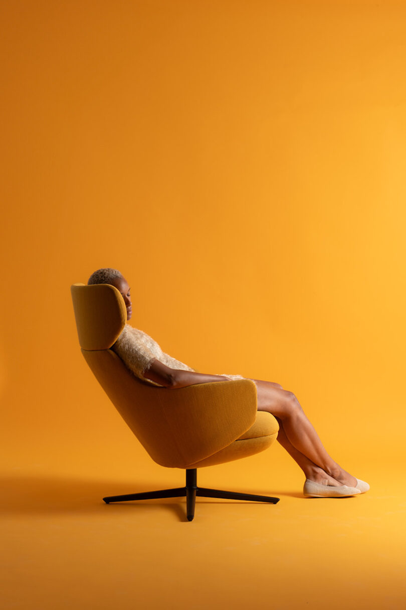 Person sitting in a modern, mustard-colored swivel chair against an orange background, wearing casual clothing and white shoes