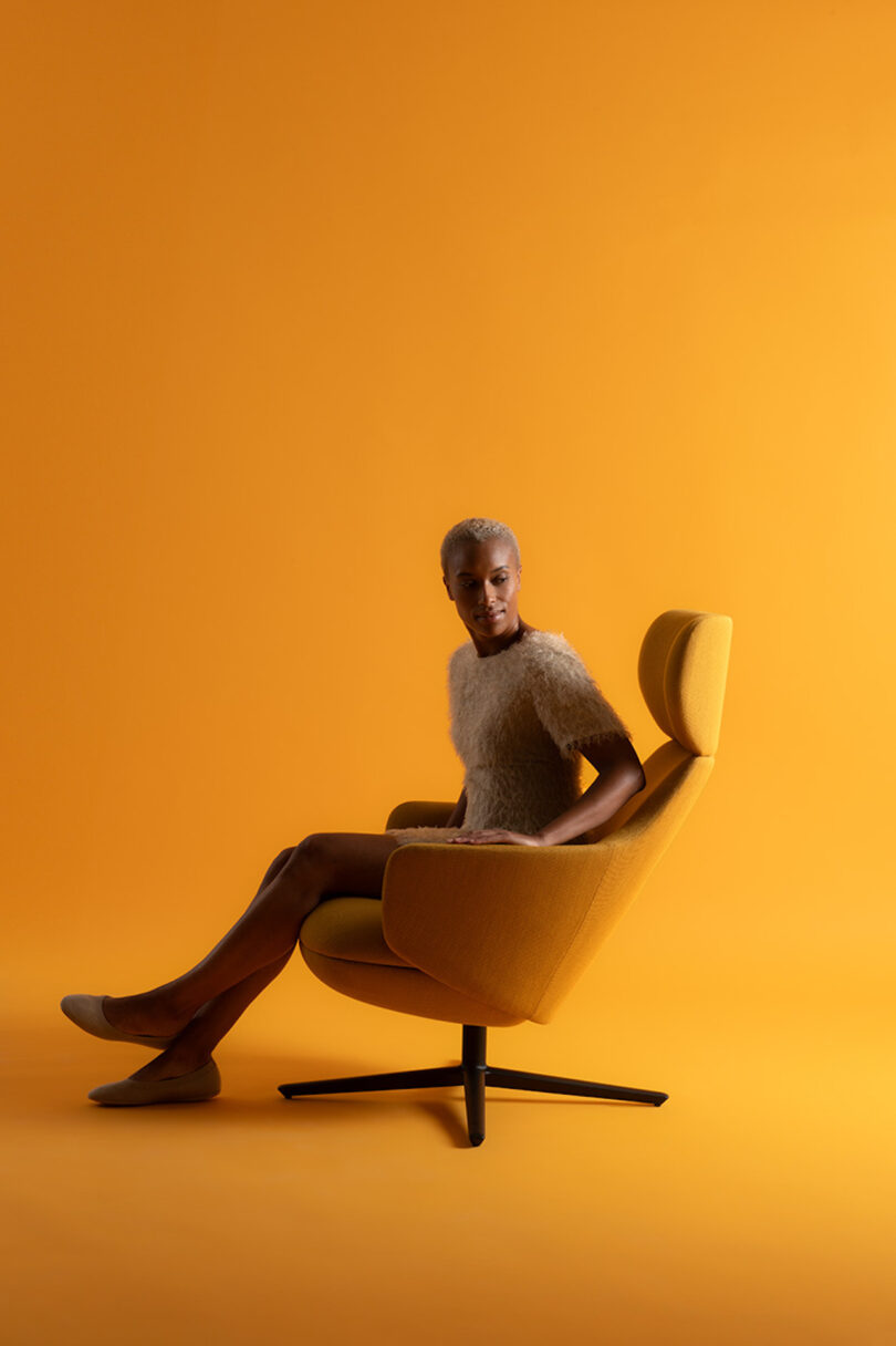 Person seated in a modern yellow chair, on a matching yellow background, dressed in a textured top and slippers, gazing forward