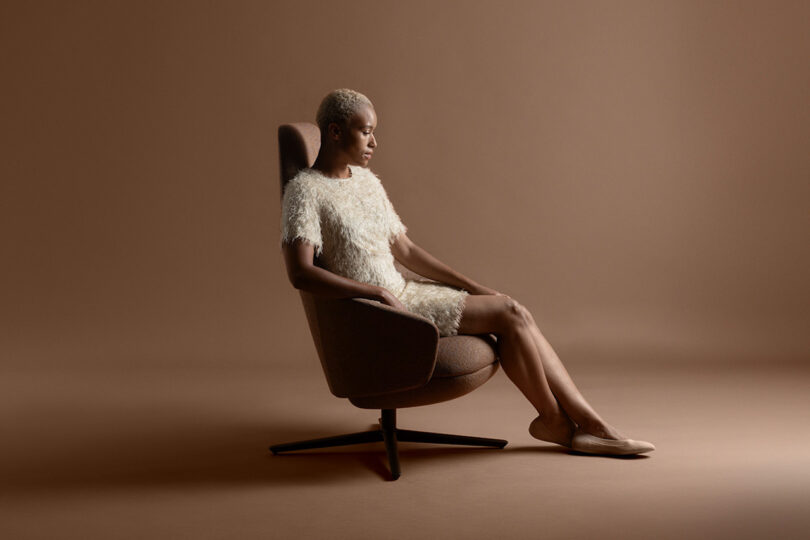 Person seated in a tall, brown swivel chair against a beige background, wearing a white textured dress and beige shoes