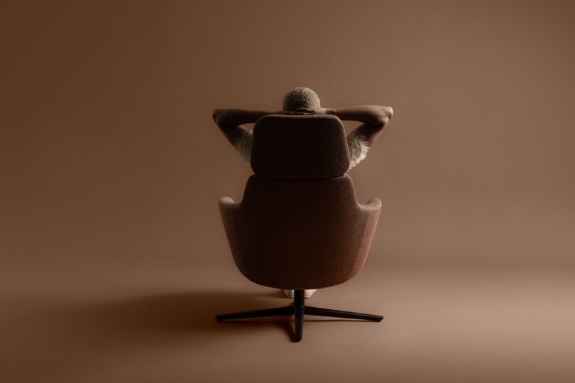 A person with short hair sits in a swivel chair, viewed from behind, resting their arms on their head against a brown background