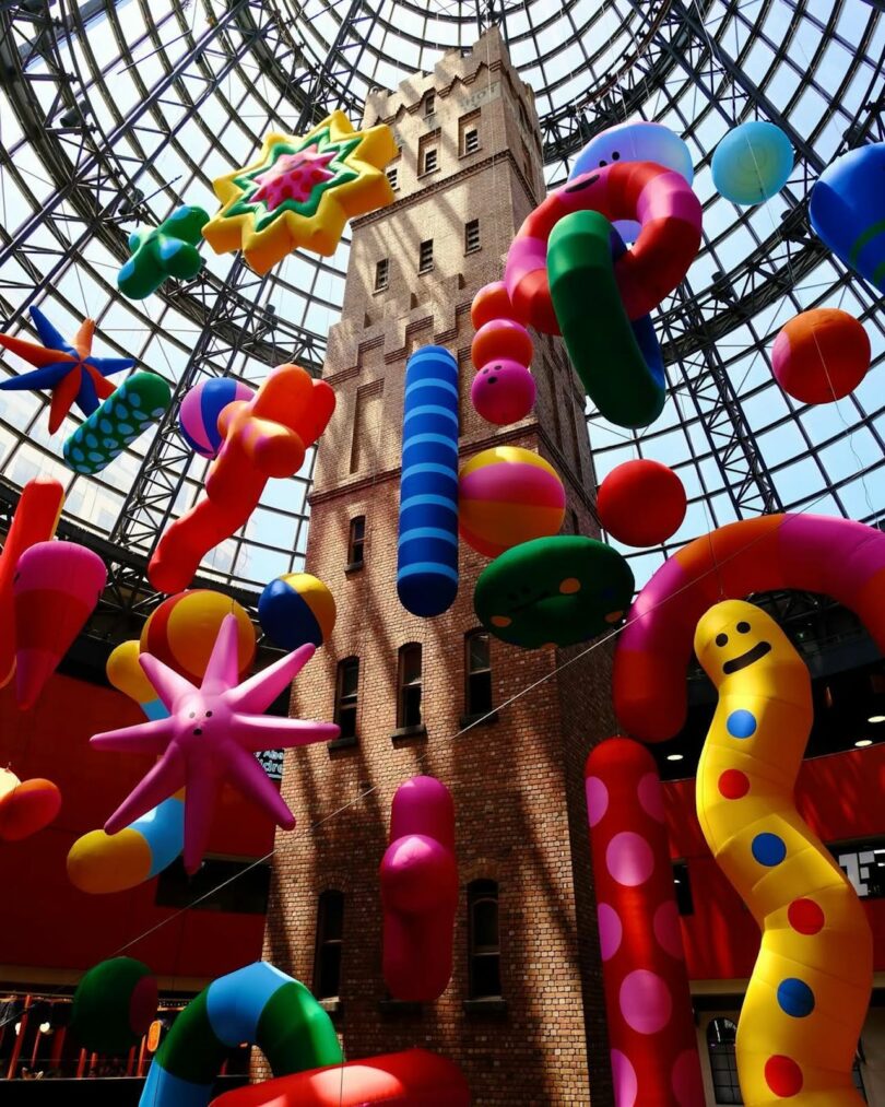 A tall brick tower is surrounded by colorful, abstract inflatables under a glass dome ceiling.