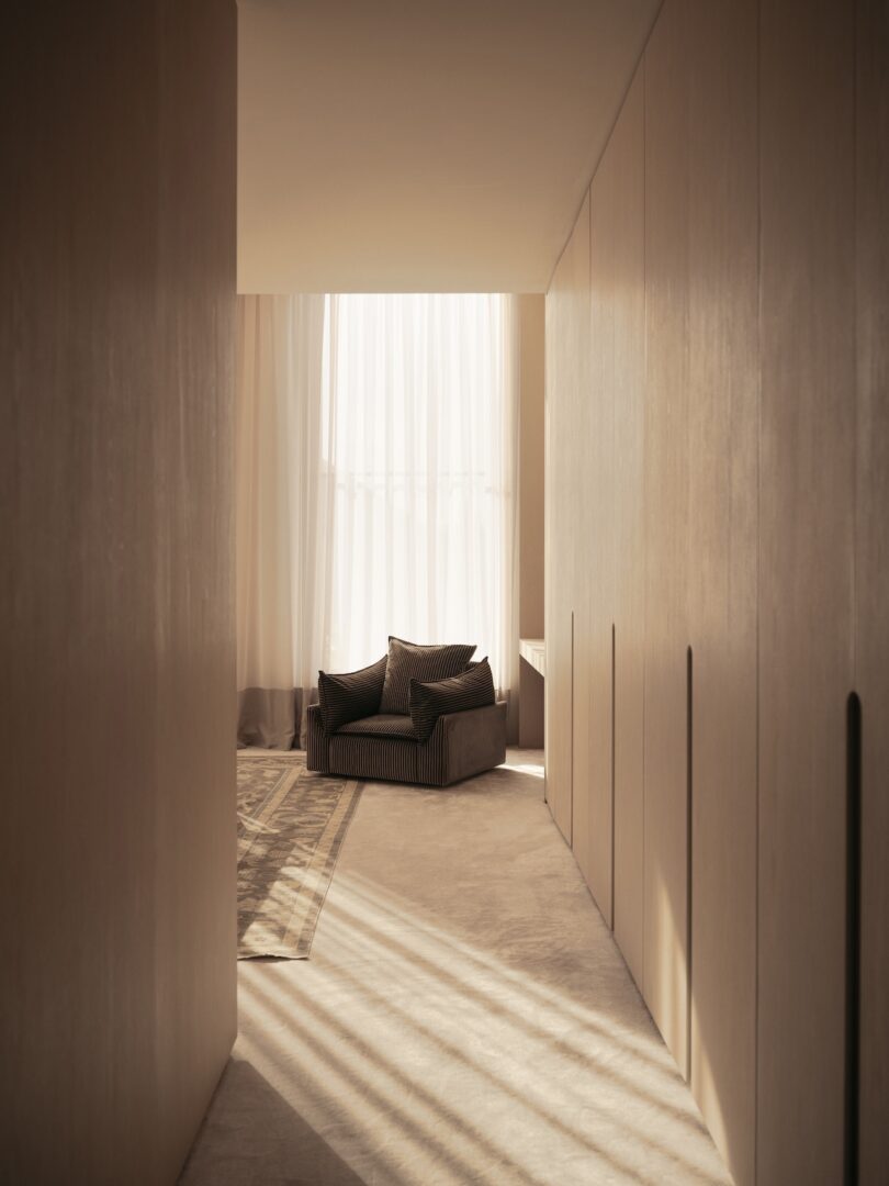 A minimalist room with a single armchair near floor-to-ceiling sheer curtains, lit by soft natural light, and flanked by light wood-paneled walls.