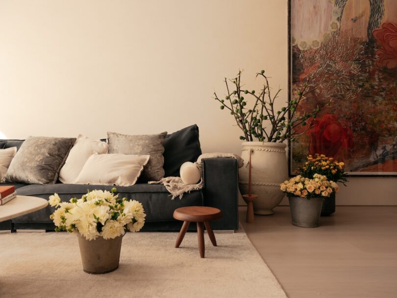 A cozy living room with a gray sofa, various cushions, a round wooden stool, potted plants, and a large abstract painting on the wall. There are flowers on the table and floor.