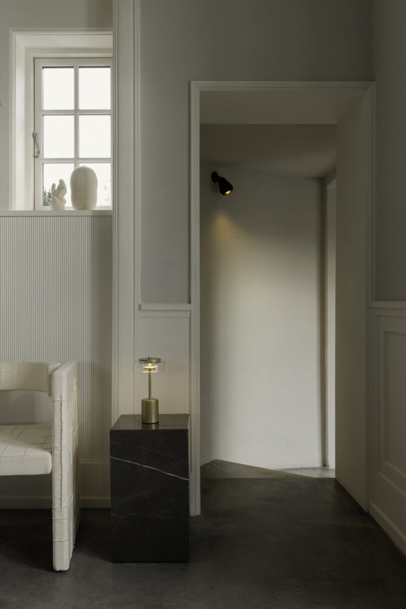 A minimalist interior features a white chair, a small table with a lamp, and a hallway illuminated by a single wall light. A small window with daylight and decor is above the chair