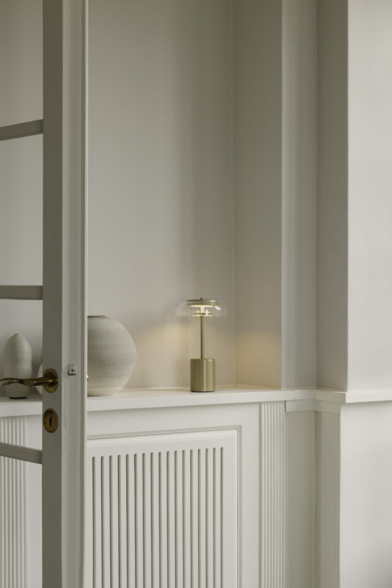 A minimalist room with light gray walls features a brass lamp and a ceramic vase placed on a white radiator cover. The corner of an open door is visible in the foreground