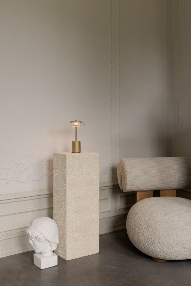 A minimalist room with a textured pedestal holding a small lamp, a bust sculpture on the floor, and a modern cylindrical chair with wooden legs on a concrete floor