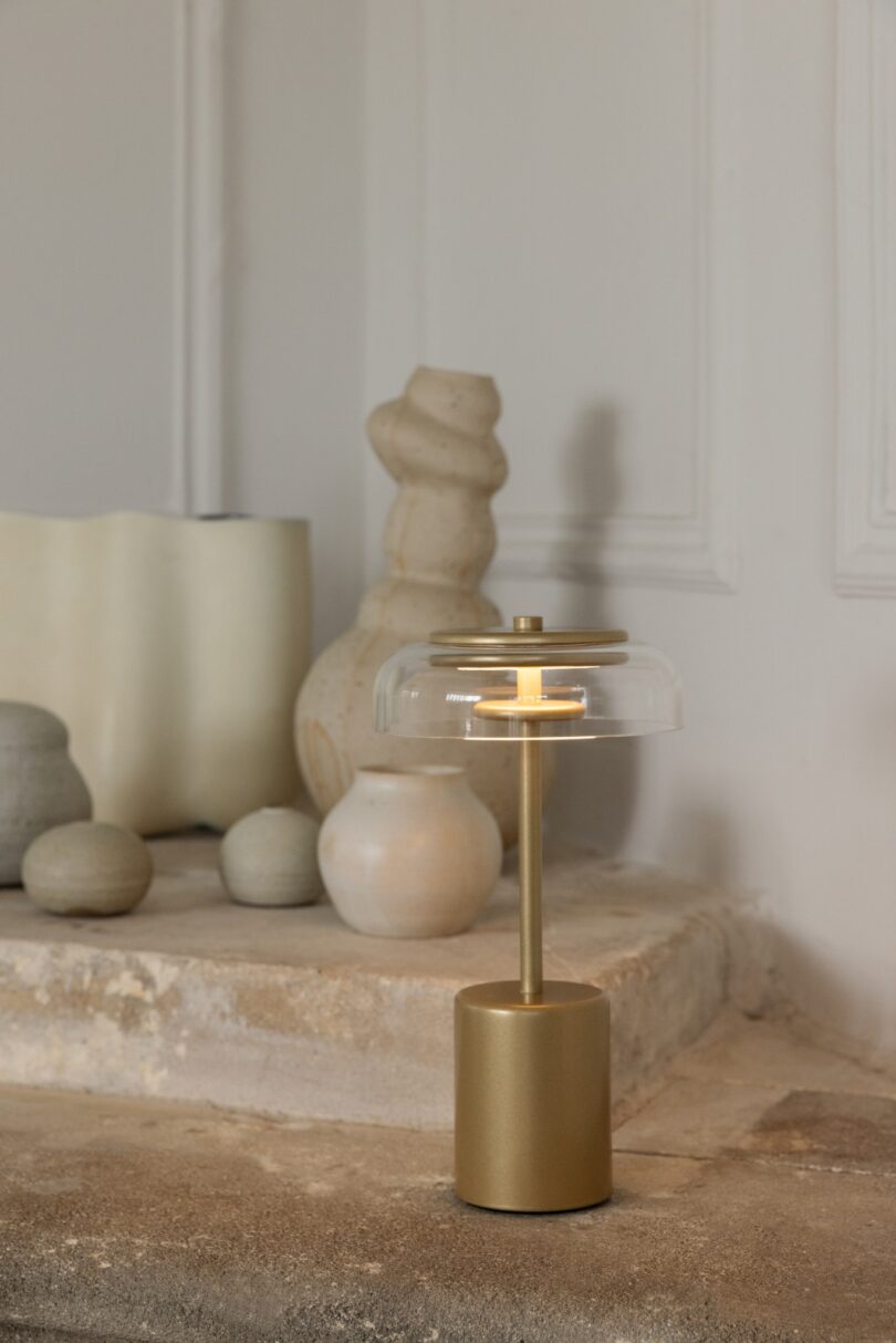 A gold table lamp with a clear glass shade sits on a stone surface, surrounded by various ceramic vases in neutral tones