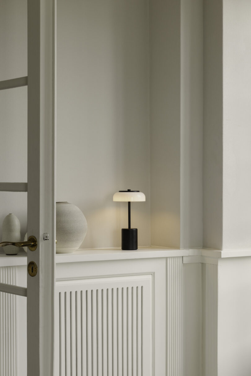 A modern black and white table lamp sits on a white cabinet next to a ceramic vase against a light gray wall in a minimalistic room