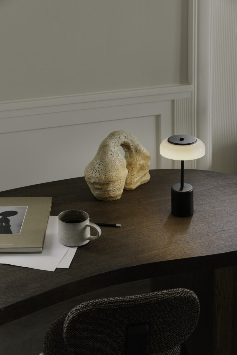 A dark wooden desk with a coffee mug, papers, a pencil, a sculpture, and a small lamp is shown. A chair is positioned in front of the desk