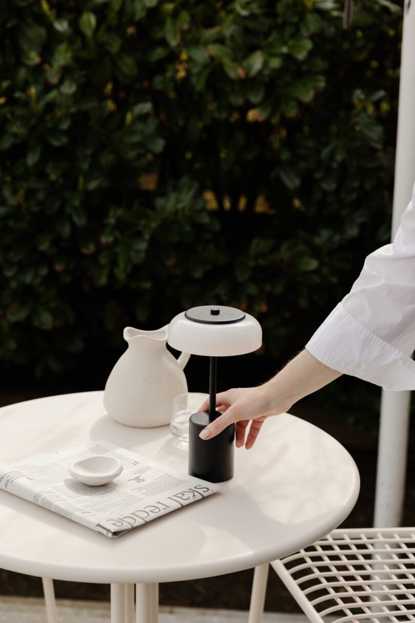 A person reaches for a small, modern lamp on a round table with a newspaper, small bowl, and a white jug, against a backdrop of greenery