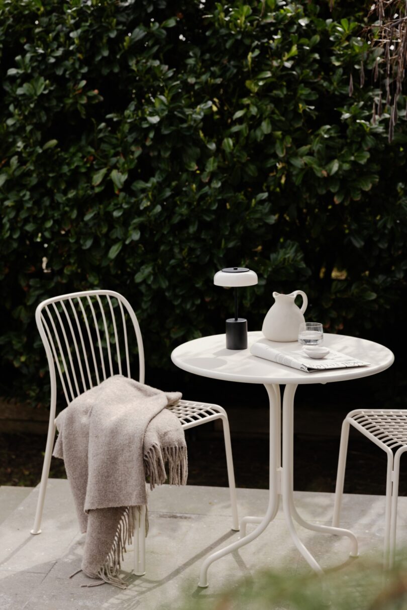 A small outdoor table with two chairs, one draped with a beige blanket. On the table are a carafe, a glass of water, a book, and a lamp. The scene is set against a backdrop of lush green bushes