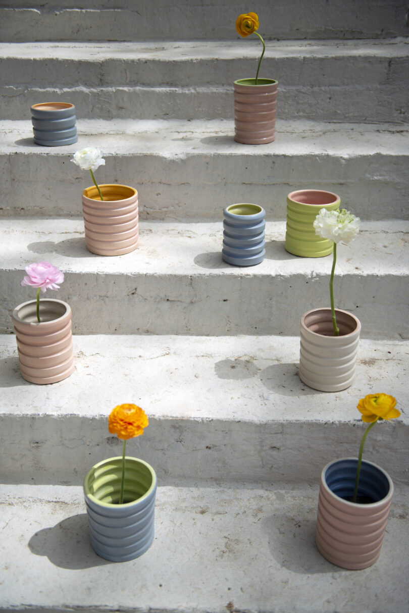 Various colored pots with single flowers are arranged on white concrete steps in a zigzag pattern