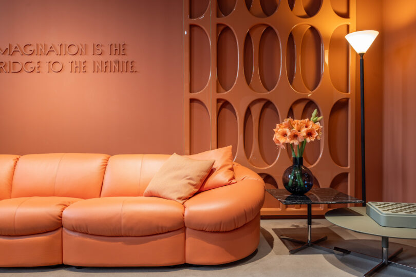 Modern living room with an orange sofa, pillows, a black table with flowers, a tall lamp, and a textured wall with the phrase "IMAGINATION IS THE BRIDGE TO THE INFINITE."