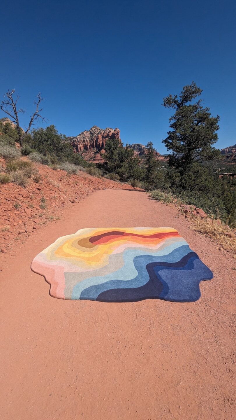 A colorful, wave-patterned rug lies on a dirt path surrounded by trees and red rock formations under a clear blue sky