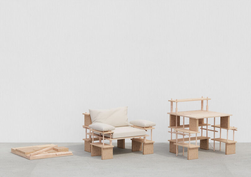 Modular wooden furniture pieces, including a chair with cushions and a shelving unit, displayed against a plain white wall. Flat packed components are placed on the floor