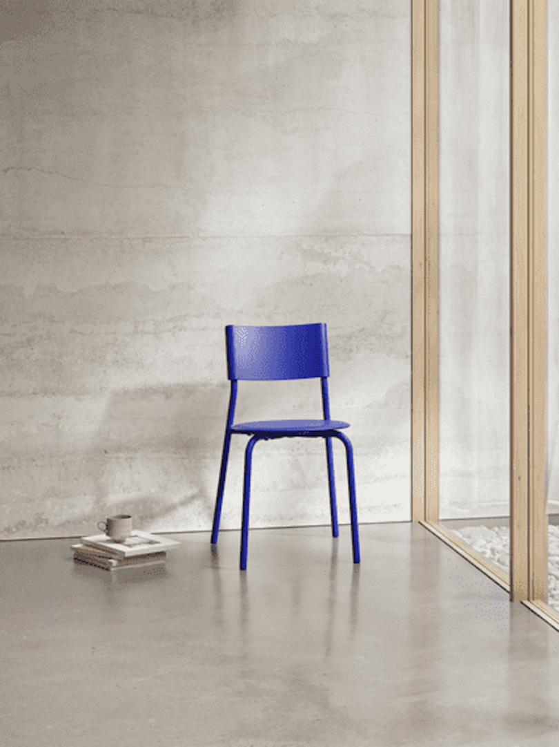 A blue chair is placed on a smooth concrete floor near a wooden framed glass wall, with a small stack of books and a cup beside it