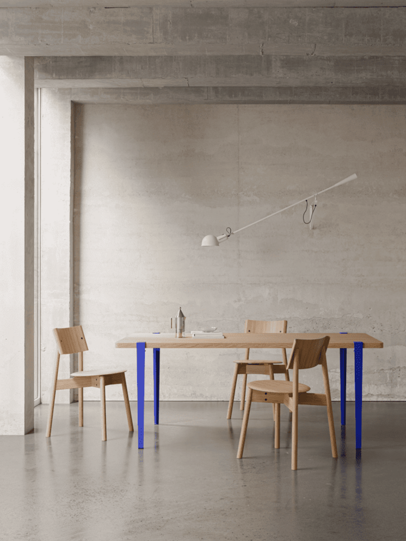 Minimalist dining area with a wooden table, three chairs, and a white wall lamp. The setting features a concrete wall and floor with blue table legs