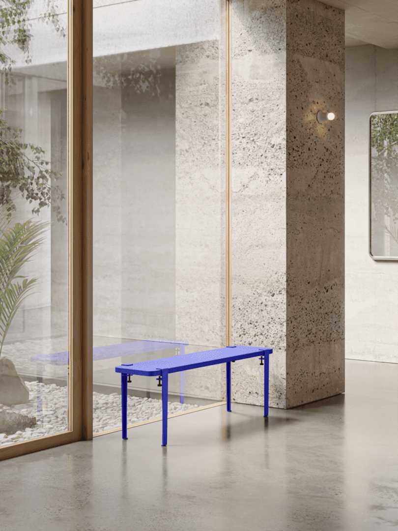 A modern, minimalist interior with a blue bench placed by large glass windows. Concrete walls and floors surround the space, complemented by a small plant outside