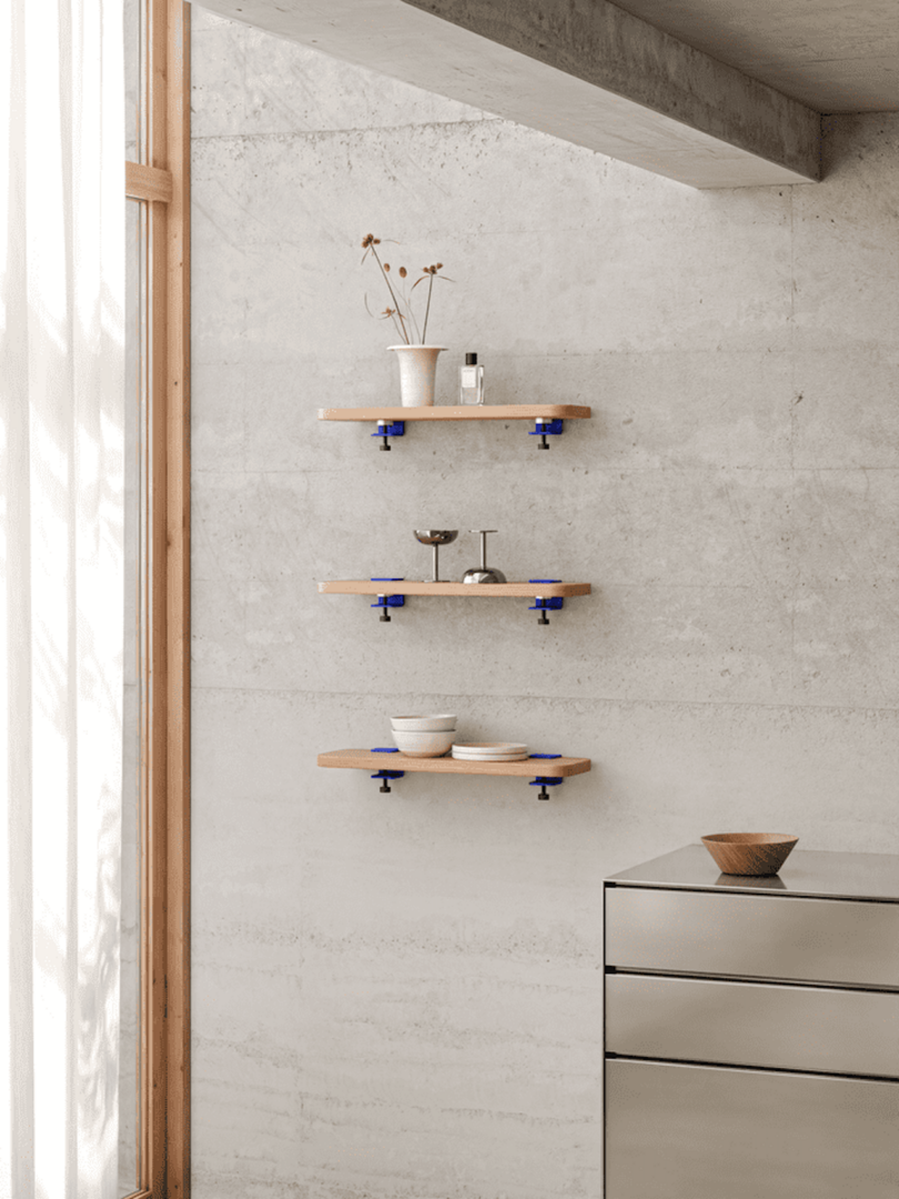 Three wooden shelves with blue brackets are mounted on a concrete wall, displaying vases, candles, and bowls. A modern cabinet with a wooden bowl on top is nearby