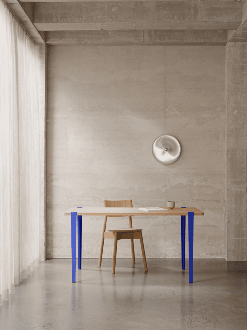 Minimalist room with a wooden table, blue legs, a chair, a mug, and a notebook. Concrete walls feature a round wall art. Soft light filters through sheer curtains