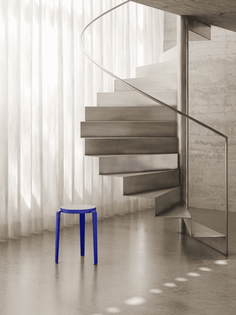 A blue stool sits on a polished floor next to a modern spiral staircase and sheer curtains