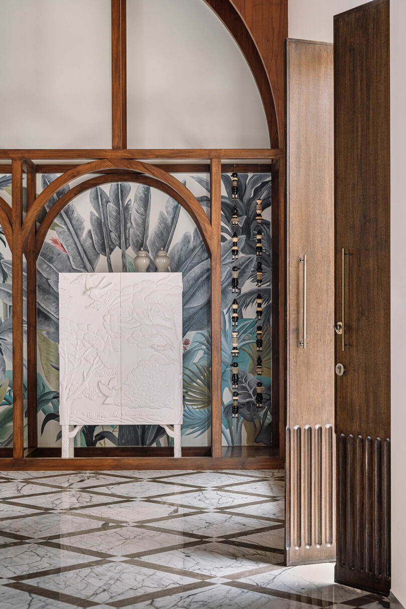 A room with a marble floor features a wooden door, wooden arch frames, and a white cabinet with decorative carvings against a backdrop of tropical-themed wallpaper.