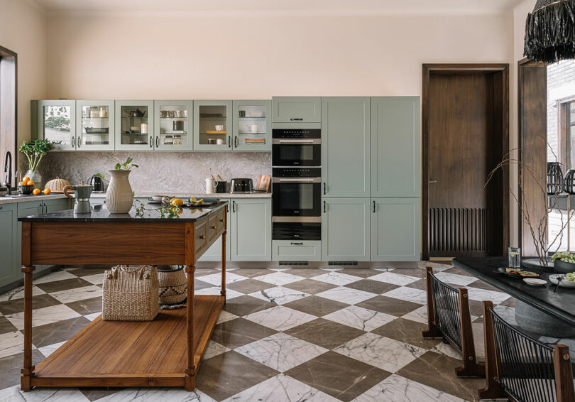 A spacious kitchen with sage green cabinets, marble countertops, a wooden island, and a checkered marble floor. The room is well-lit with a mix of natural and artificial light.