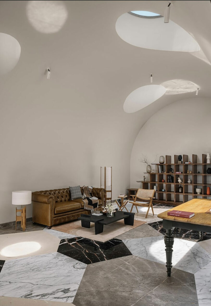 A spacious living area with a brown leather sofa, a wooden chair, a large design table, and a bookshelf against a white wall. The room has a unique ceiling with multiple skylights and geometric flooring.