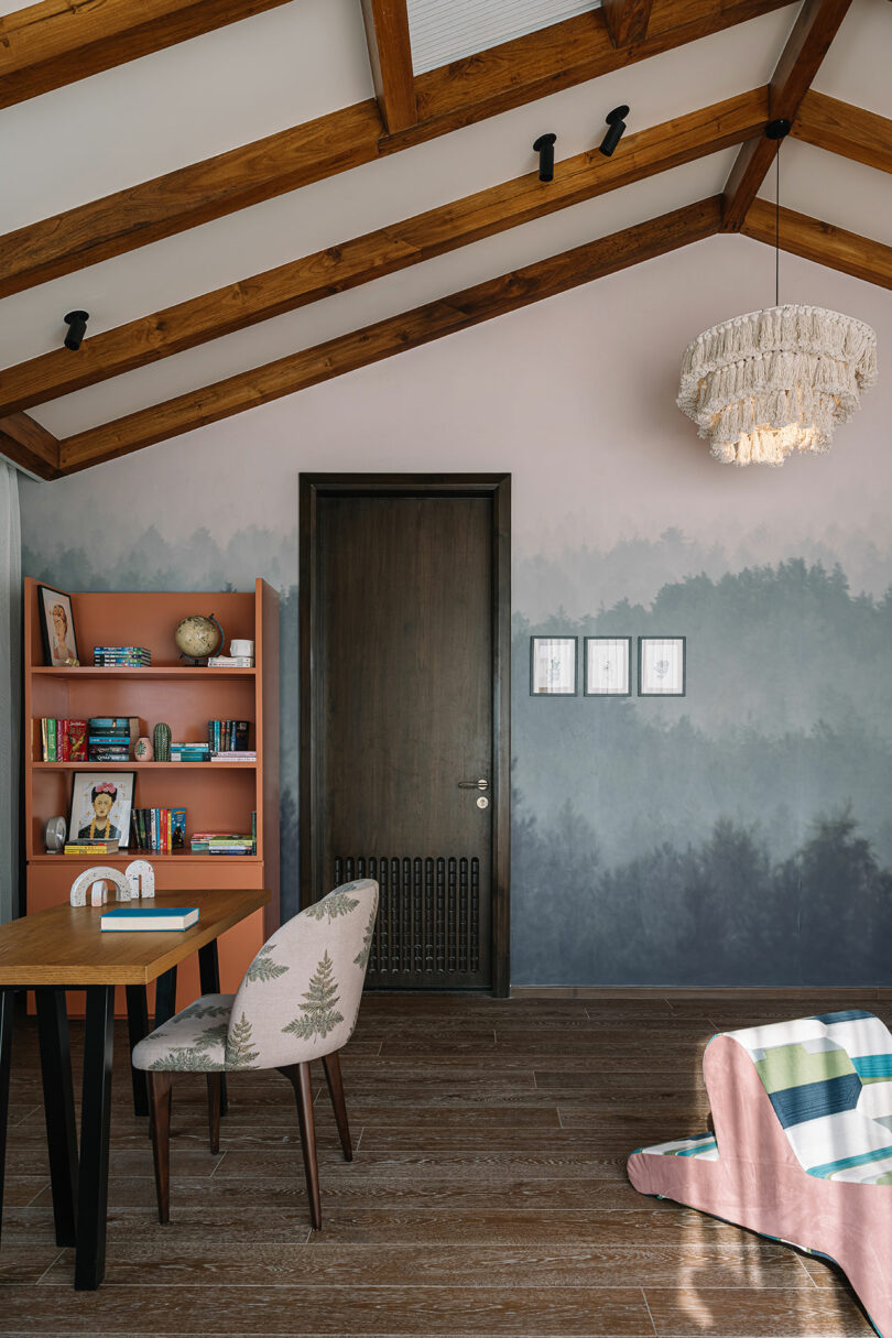 A home office with wooden beams on the ceiling, a desk with a chair, a coral bookshelf, and framed pictures on a misty forest-themed wall. A chandelier hangs from the ceiling.