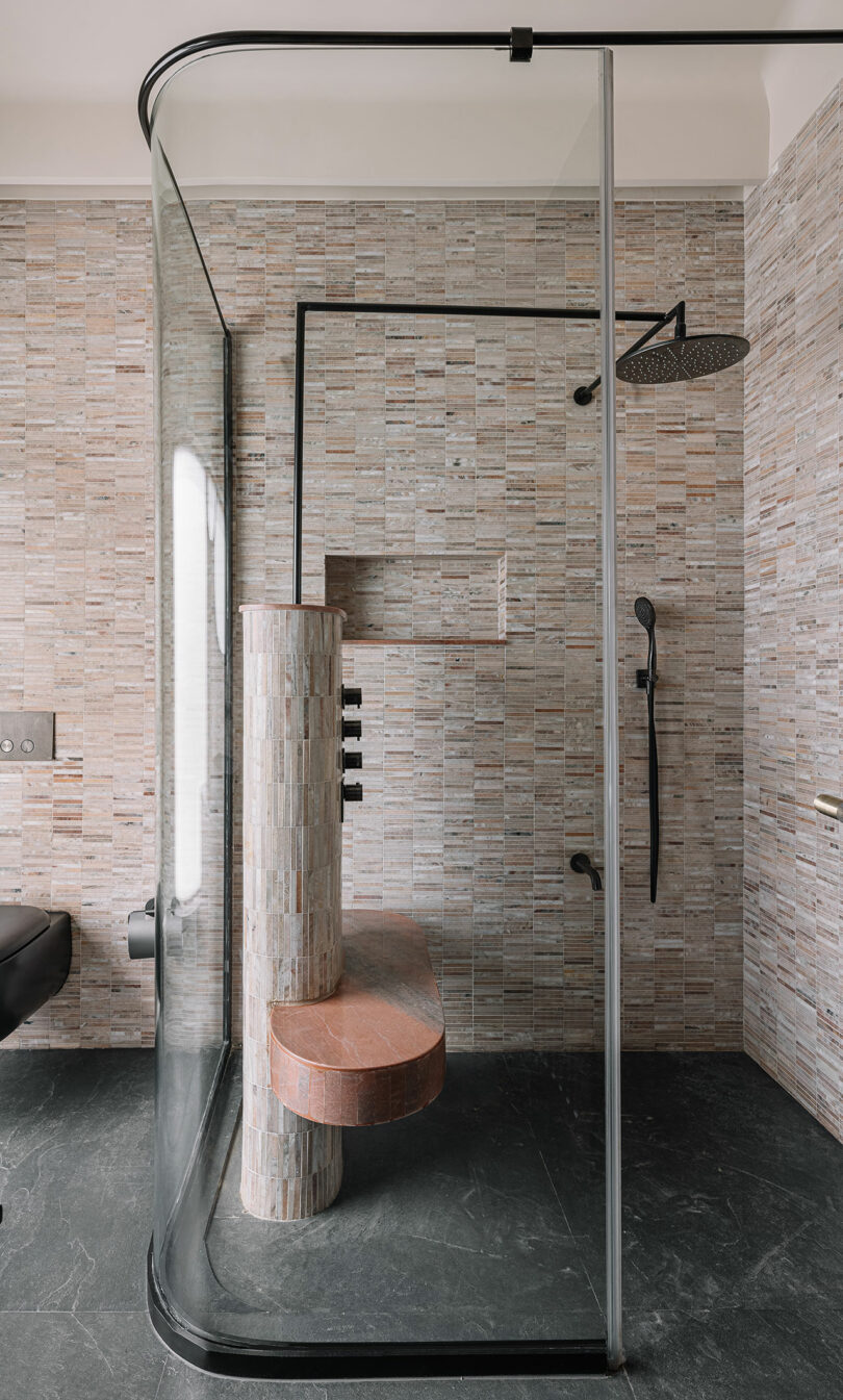 A modern bathroom features a curved glass shower enclosure with brick-patterned tiles, a round stone bench, a rainfall showerhead, and a sleek black handheld shower.