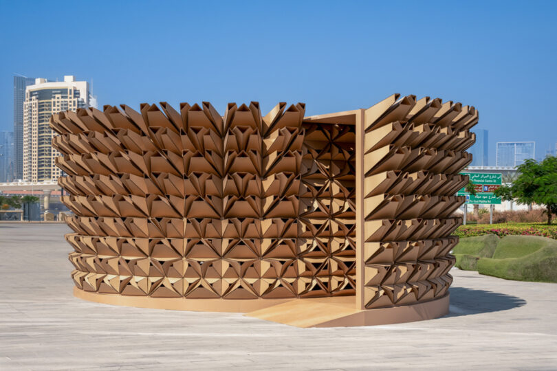 A modern, circular art installation with a textured, wooden geometric pattern on an outdoor plaza under a clear blue sky.