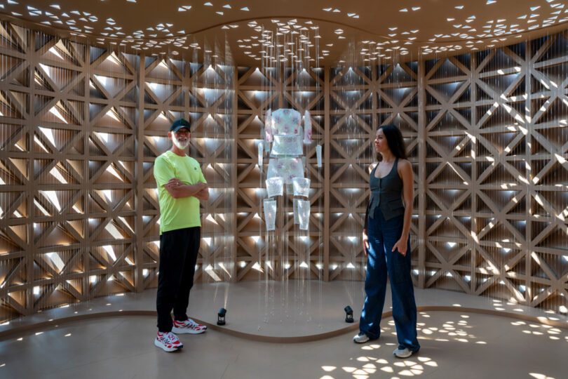 Two people stand near a transparent astronaut suit suspended in a room with geometric patterned walls and dappled light.